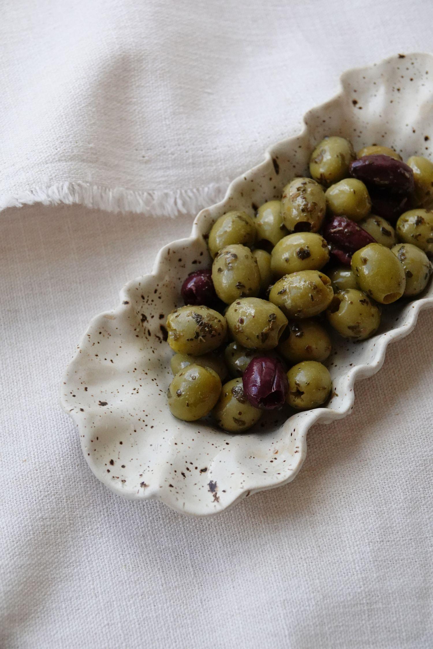 kalamata and green olives dressed in olive oil and herbs served in a ceramic oval, cream colored with brown speckled glaze and a scalloped rim. Resting on a textured white linen cloth