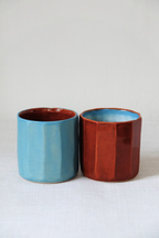 Two glazed ceramic cups with a faceted cylindrical design, one in light blue with a reddish-brown interior, and the other in reddish-brown with a light blue interior, set against a neutral light gray background.