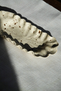 A speckled off-white ceramic dish with a wavy, scalloped rim, resting on a textured white fabric surface under direct sunlight.