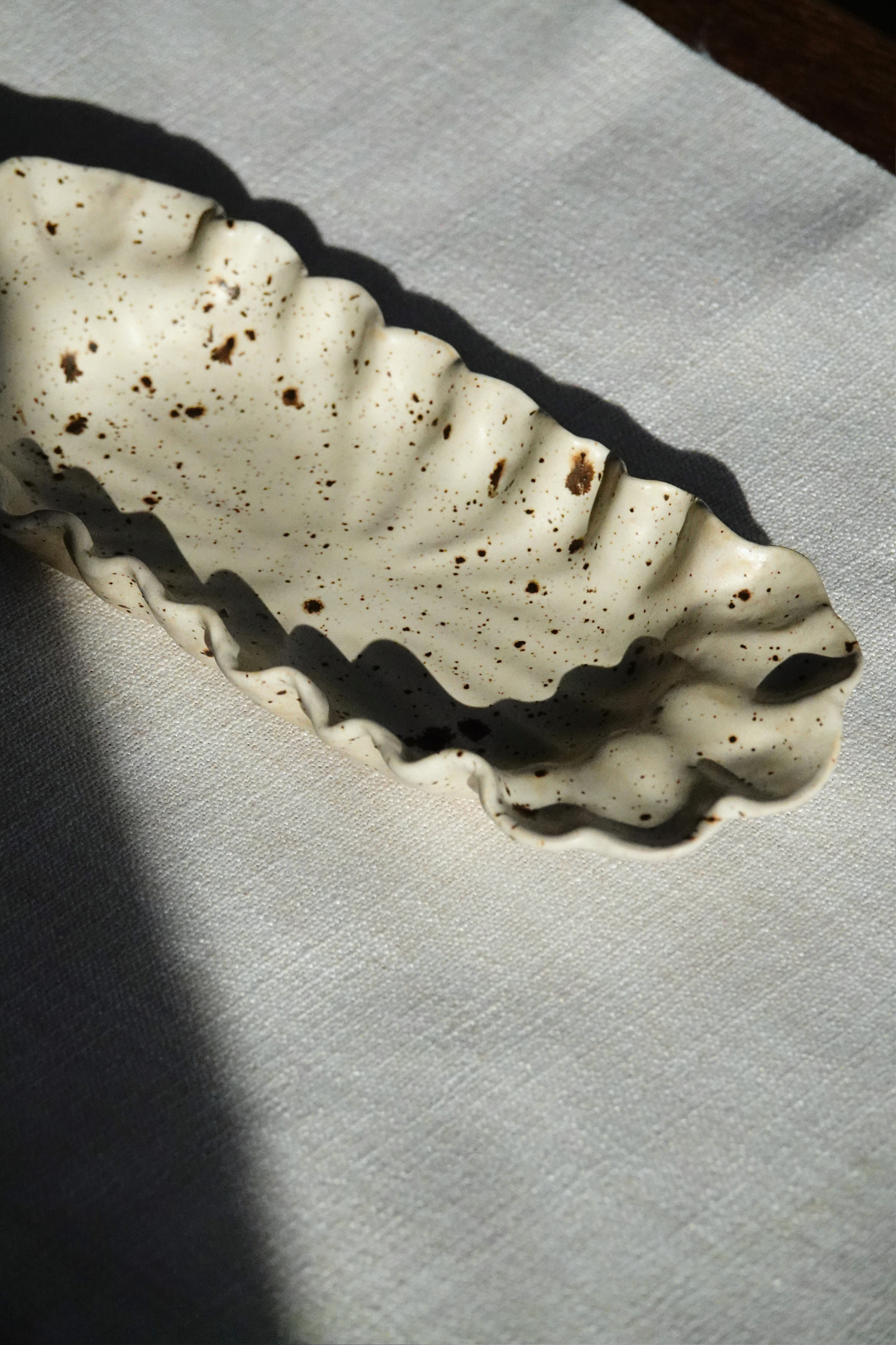 A speckled off-white ceramic dish with a wavy, scalloped rim, resting on a textured white fabric surface under direct sunlight.