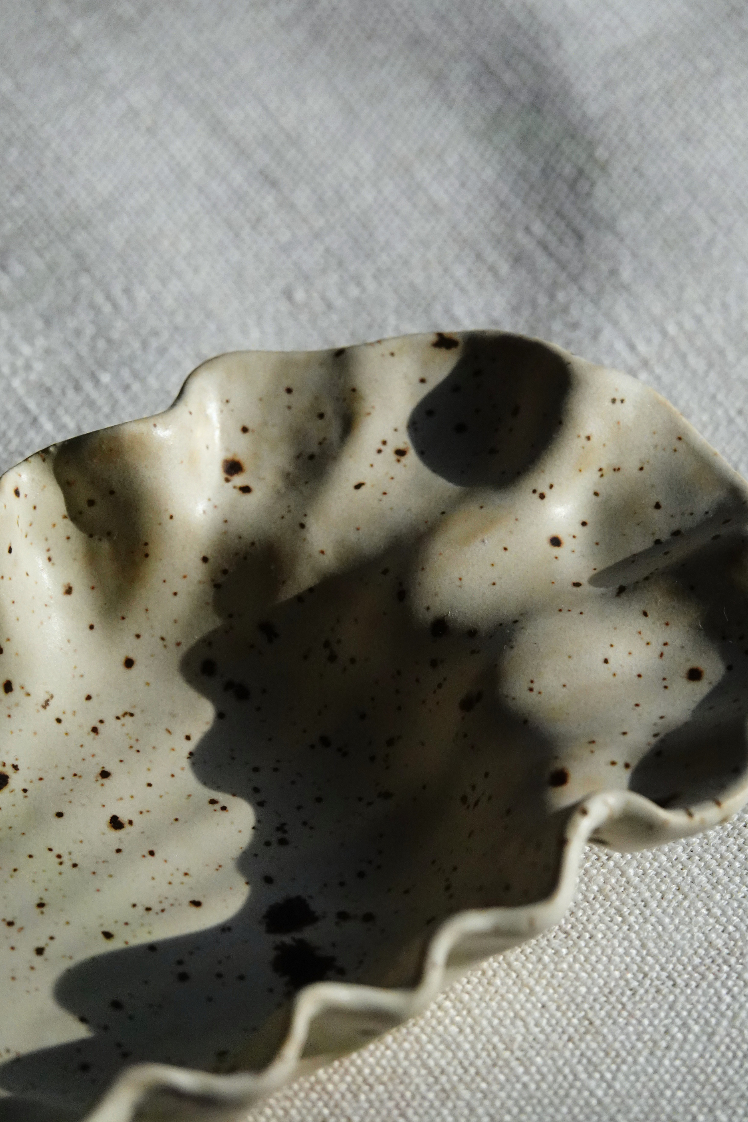A close-up of a ceramic dish with a scalloped rim, featuring a speckled cream glaze with dark brown and black flecks, set against a textured light cream fabric background with dappled shadows.