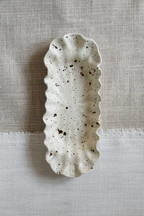 A speckled ceramic serving dish with a scalloped edge, shown from a top-down view on a textured linen fabric background.