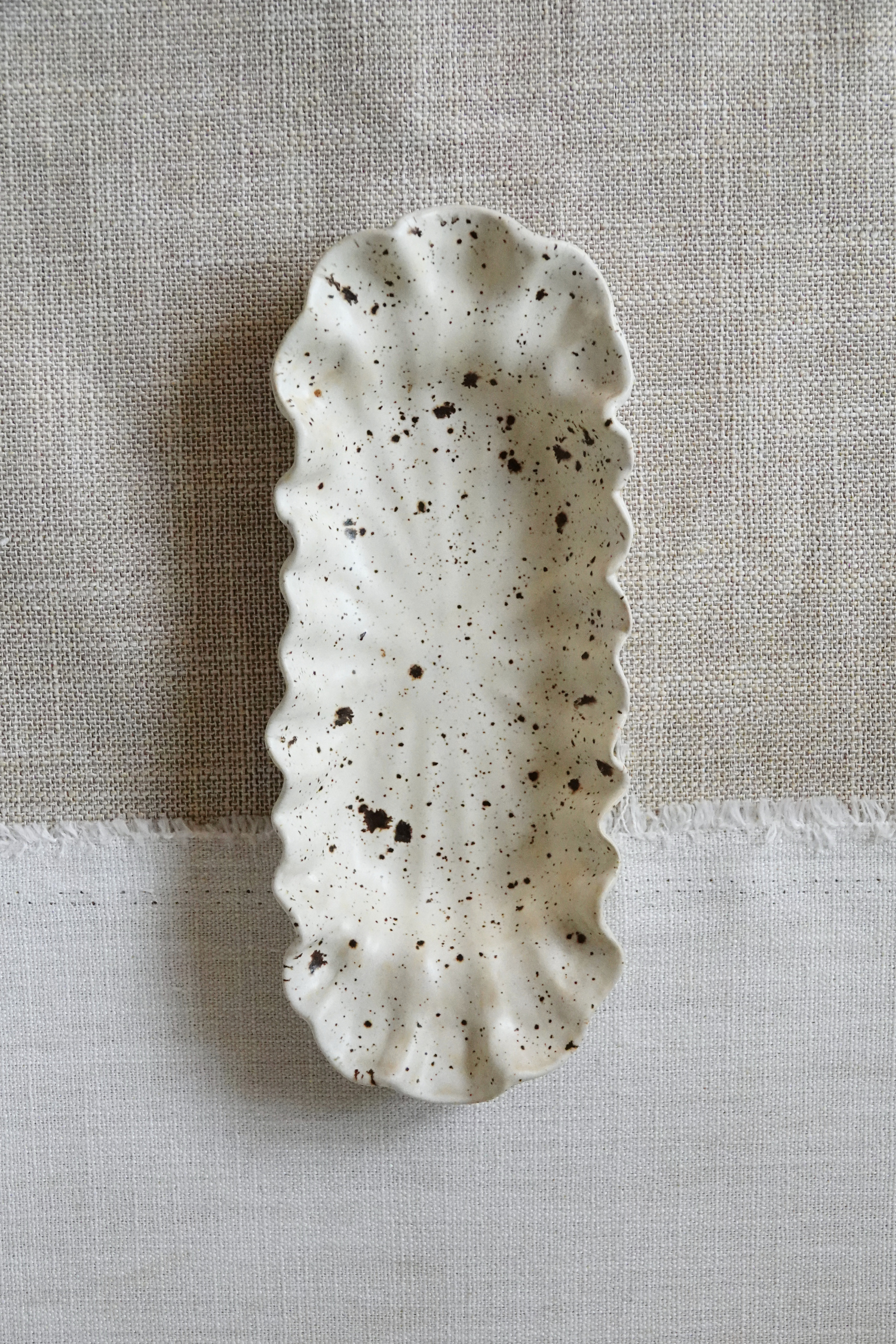 A speckled ceramic serving dish with a scalloped edge, shown from a top-down view on a textured linen fabric background.