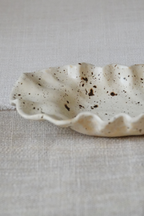 A small ceramic dish with a scalloped rim and dark brown speckles on an off-white base, resting on a textured beige linen surface.