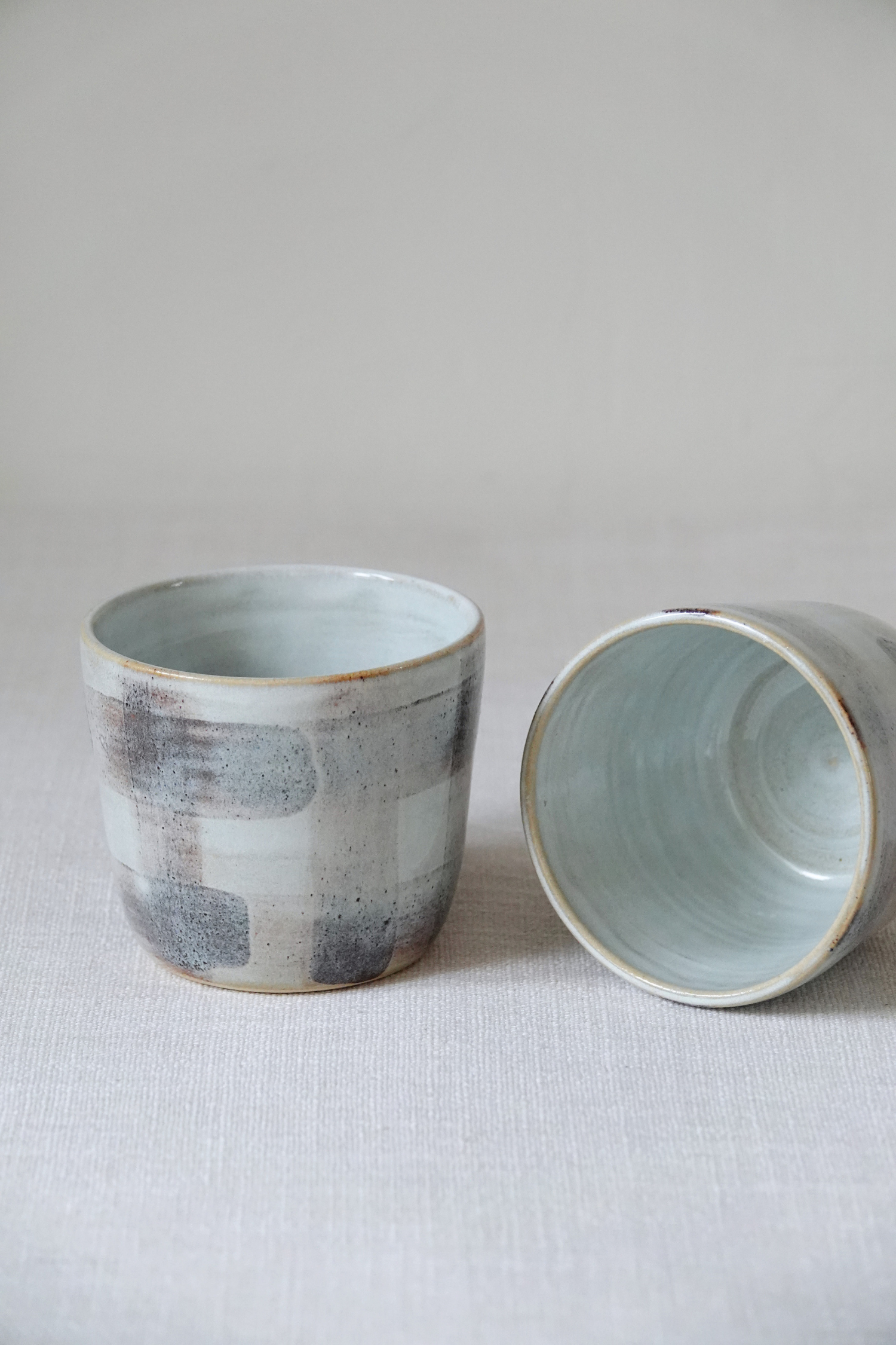 Two handmade ceramic cups with a textured, rustic glaze in brown, white, and light blue-green, shown on a neutral fabric surface.